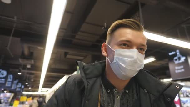 Muotokuva nuoresta miehestä, jolla on lääketieteellinen kasvonaamio kävelemässä supermarketissa. Mies, jolla on virukselta suojaava naamio. Ostoksilla kaupassa sepelvaltimotautipandemian aikana. Terveyden käsite. COVID-19 - Materiaali, video