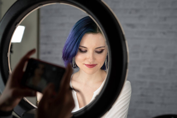 Nuori kaunis tyttö ammatillinen meikki, siniset hiukset ja täydellinen iho istuu studiossa edessä rengas valo lamppu poseeraa sosiaalisen median. Kauneusblogi. Kopioi tila
. - Valokuva, kuva