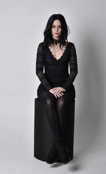 Portret gotyckiej dziewczyny z ciemnymi włosami w czarnej koronkowej sukience i butach. Pełna długość pozycji siedzącej na tle studia. - Zdjęcie, obraz
