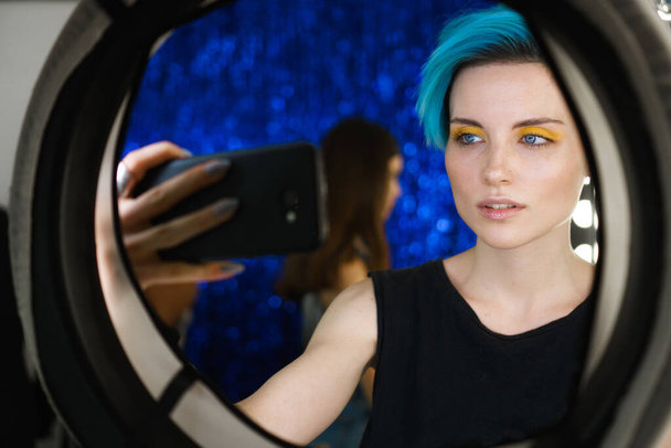 dziewczyna z niebieskimi włosami i makijaż żółte oko zrobić selfie na telefonie w studiu piękności przed świetle pierścienia w mediach społecznościowych. - Zdjęcie, obraz