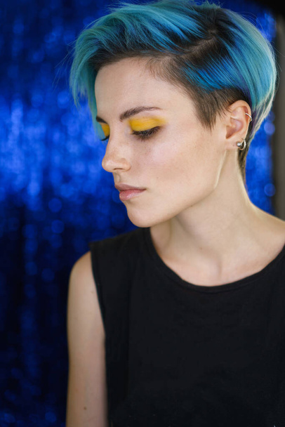 Retrato de una chica con pelo azul y maquillaje de ojos amarillos en un salón de belleza frente a una luz de anillo sobre fondo azul brillante. Disparo vertical. Ojos cerrados
 - Foto, Imagen