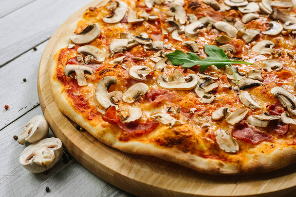 Maukas pizza Caprizzioza tomaattikastikkeella, kinkulla ja herkkusienillä valkoisella puupohjalla
 - Valokuva, kuva