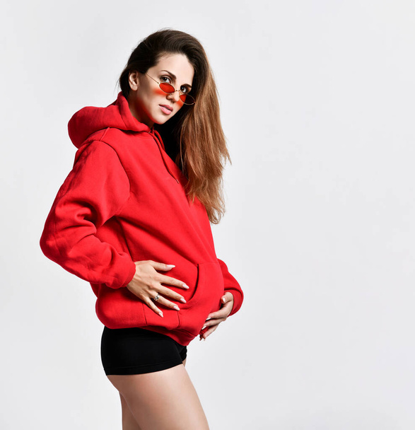 Pregnant female in black sport shorts, red hoody. She put hand on her belly, smiling, posing sideways isolated on white. Close up - Valokuva, kuva
