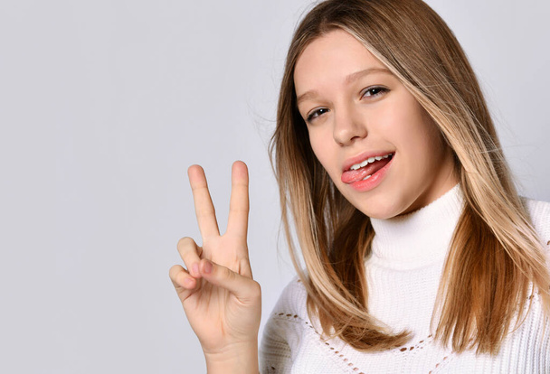 Блондинка дівчина-підліток в светрі і браслеті. Вона показує язик і руку перемоги, позує ізольовано на білому. крупним планом
 - Фото, зображення