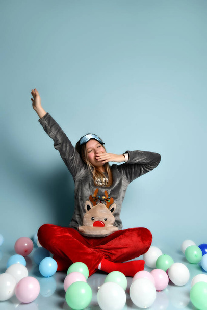 Teen girl in reindeer pajamas, red socks, sleep mask. Yawning, closed eyes, sitting on floor, balloons, blue background. Close up - Photo, Image