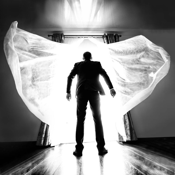 Ένας άντρας με ένα επαγγελματικό κοστούμι μπροστά σε ένα λαμπρό φως. Καλλιτεχνική υπερέκθεση ενός άνδρα στο κατώφλι ενός καινούργιου. Ασπρόμαυρη φωτογραφία ενός άνδρα από πίσω μπροστά από ένα πολύ φωτεινό φως - Φωτογραφία, εικόνα