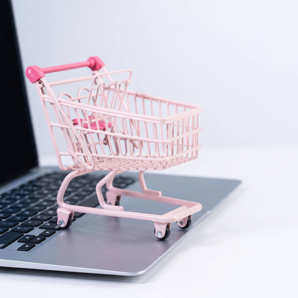 Online shopping. Mini empty pink shop cart trolley over a laptop computer on white table background, buying at home concept, close up - Photo, image