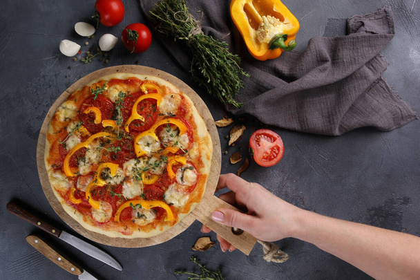 Italian cuisine. Pizza with mozzarella, yellow peppers, tomatoes and thyme on a wooden board on a grey background. Vegetable pizza with hands. Rustic. Background image, copy space - Fotó, kép