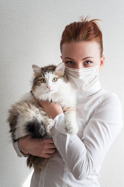 Portrait of a girl in a medical mask. She is holding a pet cat. Close u - Photo, Image
