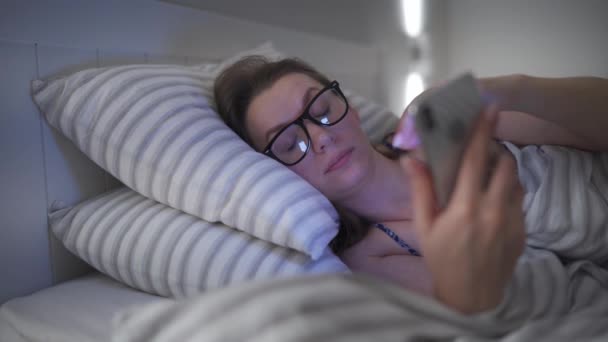 Woman in glasses using smartphone while lying in bed. She rubs her eyes, because she is tired and sleepy. Concept of increased stress and fatigue. - Materiaali, video