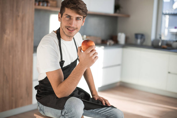 attractive man with an apple sitting in a home kitchen - Foto, Imagem