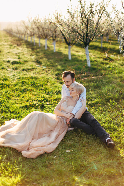 Happy pleasant romantic Caucasian couple sitting on the green grass and hugging, in beautiful blooming garden, enjoying spring nature. Tenderness and love concept - Photo, Image