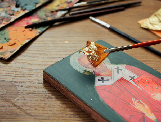 Altın yapraklı el fırçası. Sanatçı ikona altın yaprak uygular. Aziz Niklas ikonunun kopyası. Sanatçının çalışma süreci. Arkaplanda fırçalar ve boyalar. - Fotoğraf, Görsel