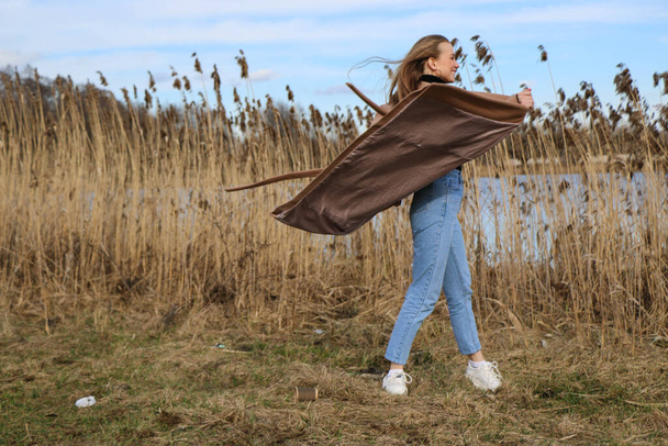 Outdoors lifestyle fashion portrait of happy stunning blonde girl. Beautiful smile. Long light hair. Wearing stylish coat. Joyful and cheerful woman. walk on a natural landscape, near a dry reed and a lake on a sunny day. - Foto, Imagem
