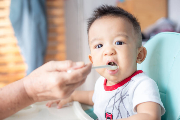 Asiático bebé niño sentado en silla de bebé tratar de comer mezcla de alimentos en casa - Foto, imagen