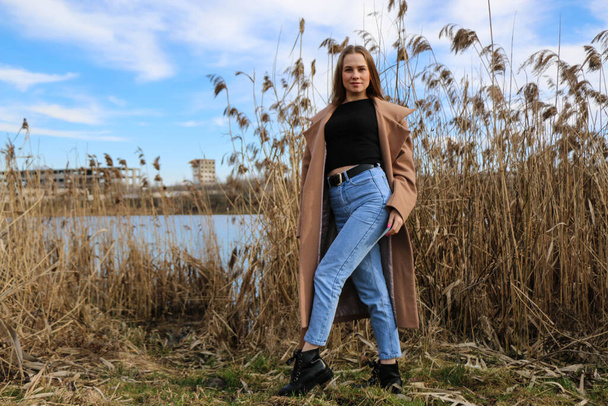 Outdoors lifestyle fashion portrait of happy stunning blonde girl. Beautiful smile. Long light hair. Wearing stylish coat. Joyful and cheerful woman. walk on a natural landscape, near a dry reed and a lake on a sunny day. - Zdjęcie, obraz