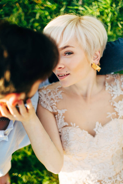 Top view portrait of charming lovely blond woman bride, looking at camera, while lying on the knees of her handsome loving man, outdoors in beautiful garden with green grass. Spring love photo - Foto, Imagem