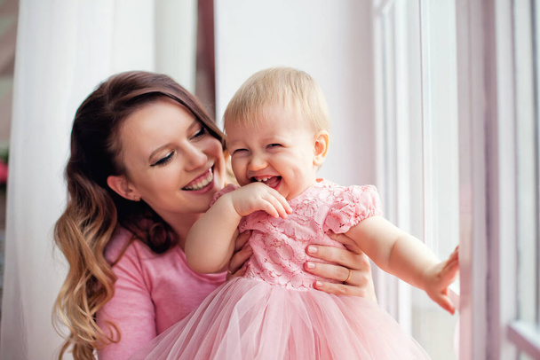 Η μητέρα παίζει με την κόρη της στο παράθυρο με ροζ φορέματα. Ημέρα της μητέρας. - Φωτογραφία, εικόνα