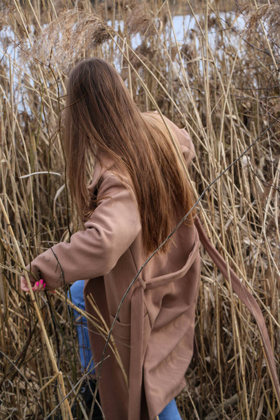Pretty young woman posing at camera outdoor. Bulrush background. Outdoor. Close up. On a sunny day near the dry reed and blue lake. Beautiful blonde with long hair In a brown coat - Φωτογραφία, εικόνα
