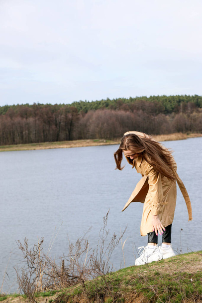 Outdoors lifestyle fashion portrait of happy stunning blonde girl. Beautiful smile. Long light hair. Wearing stylish coat. Joyful and cheerful woman. walk on a natural landscape, near a dry reed and a lake on a sunny day. - Foto, Bild