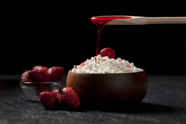 adding strawberry jam to fresh white cottage cheese with berries in a wooden plate against a dark background, cream pour on strawberries in cottage cheese, close-up - Photo, Image