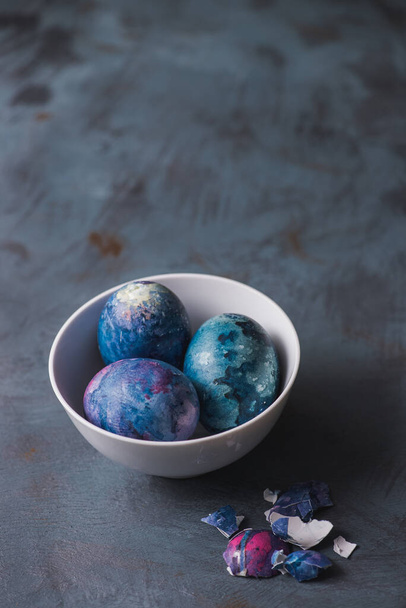 Huevos de Pascua garabateados a mano en colores coloridos en un tazón blanco sobre un fondo de textura azul
. - Foto, Imagen