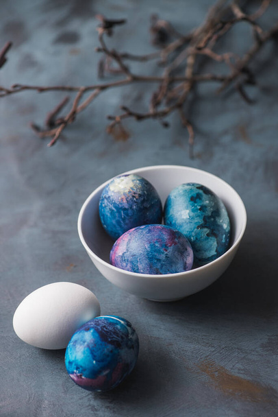 Easter composition. Easter painted colorful eggs, white bowl and branch on a blue textured background. - 写真・画像