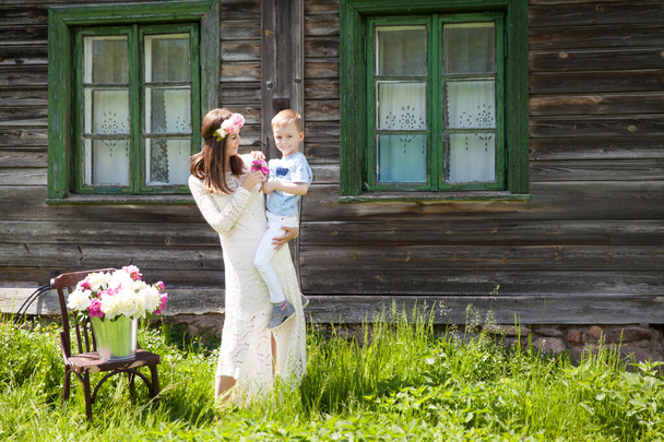 Beautiful young woman with her son near old wooden house with green windows with vintage curtains - Фото, изображение
