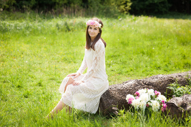 Beautiful young woman in a floral wreath sitting on a big old log in summer garden - Foto, immagini