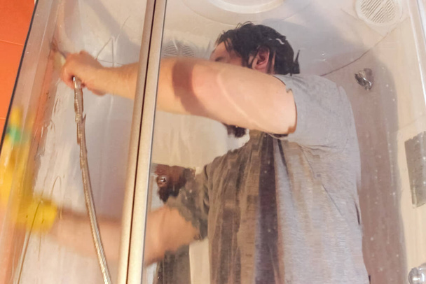 Санитарная чистка душевой кабины для безопасности дома во время коронавирус.Мужчина в серой футболке и бороде, одетый в одноразовую маску, дезинфицирует ванную комнату желтыми химическими перчатками и чистящим средством
 - Фото, изображение