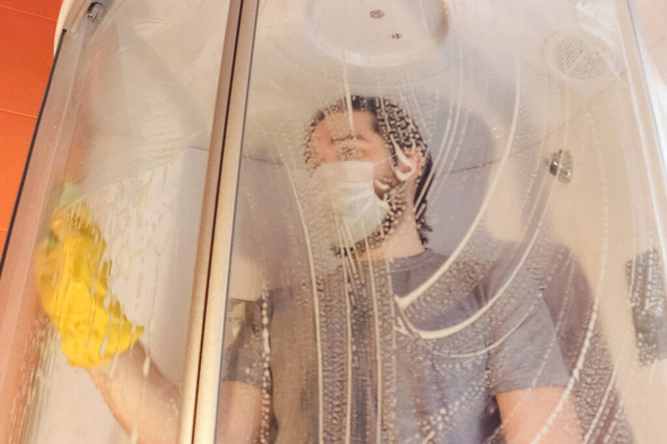 Sanitary cleaning of the shower cabin for home safety during coronavirus.A man in a gray t-shirt and beard, wearing a disposable mask, disinfects the bathroom using yellow chemical gloves and cleaning agent - Photo, Image