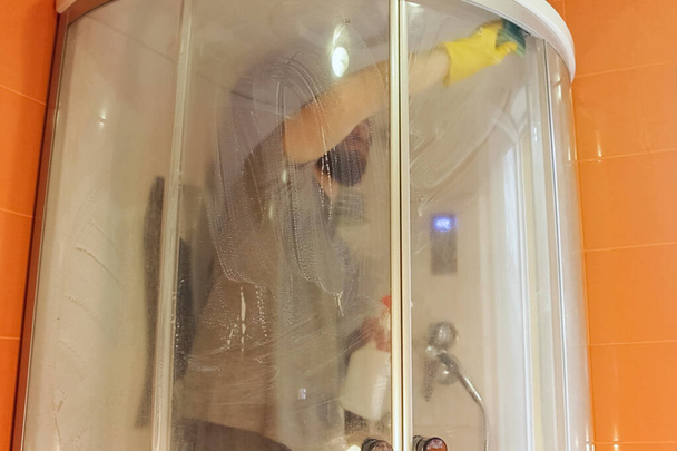 Υγειονομικός καθαρισμός καμπίνας ντους για την ασφάλεια κατά τη διάρκεια coronavirus.man σε γκρι t-shirt και γενειάδα, απολυμαίνει το μπάνιο με κίτρινα χημικά γάντια και καθαρό παράγοντα. - Φωτογραφία, εικόνα