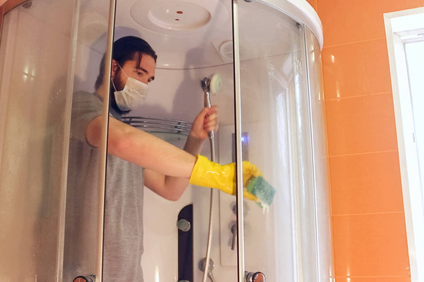 Υγειονομικός καθαρισμός της καμπίνας ντους για την ασφάλεια στο σπίτι κατά τη διάρκεια coronavirus. Ένας άντρας με γκρι μπλουζάκι και γενειάδα, φορώντας μάσκα μιας χρήσης, απολυμαίνει το μπάνιο με κίτρινα χημικά γάντια και καθαριστικό. - Φωτογραφία, εικόνα