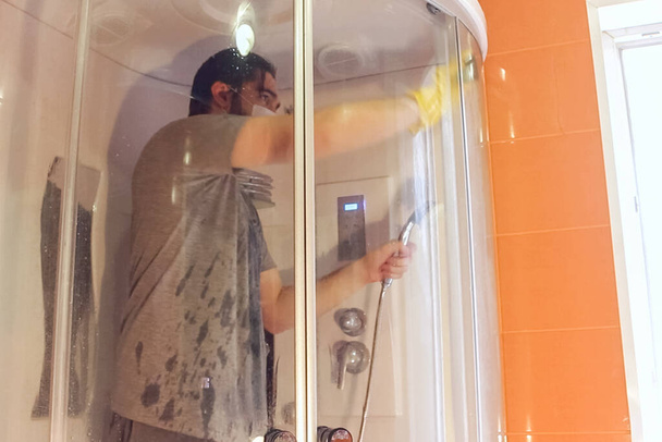 Sanitární čištění sprchovací kabiny pro domácí bezpečnost během koronaviru.Muž v šedém tričku a vousu s jednorázovou maskou dezinfikuje koupelnu pomocí žlutých chemických rukavic a čisticího prostředku - Fotografie, Obrázek