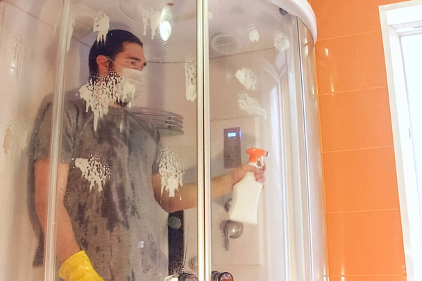Sanitäre Reinigung der Duschkabine zur Sicherheit der Wohnung bei Coronaviren. Ein Mann in grauem T-Shirt und Bart, der eine Einmalmaske trägt, desinfiziert das Badezimmer mit gelben Chemiehandschuhen und Reinigungsmittel - Foto, Bild