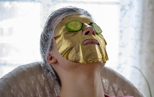 A young man with a gold cosmetic mask on his face and cucumber slices on his eyes. Home Spa, beauty treatments. - Photo, Image