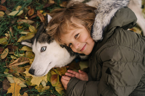 kleine jongen met krullend haar knuffelen siberische husky hond - Foto, afbeelding