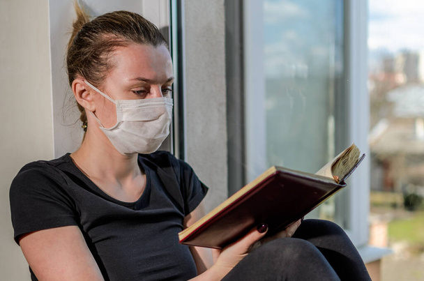 A young girl in a medical mask sits on a windowsill near a window and reads a book - Фото, изображение