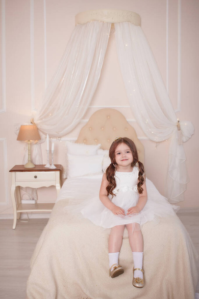 The child, the girl sits on a bed in a child's beige room, she is dressed in a white dress, and on her head is a caron. Little Princess. - Foto, afbeelding