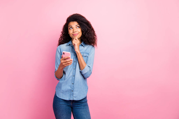 Portret van geïnspireerde sociale media verslaafd afro-Amerikaanse meisje blogger gebruik mobiele telefoon denken gedachten plan wat post in blog dragen spijkerbroek shirt geïsoleerd over roze kleur achtergrond - Foto, afbeelding