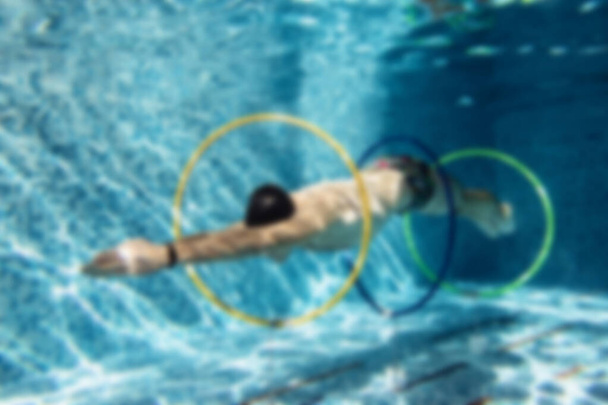 Male trainer in a cap and swimming goggles doing exercises under the water with rings in blur - Photo, Image