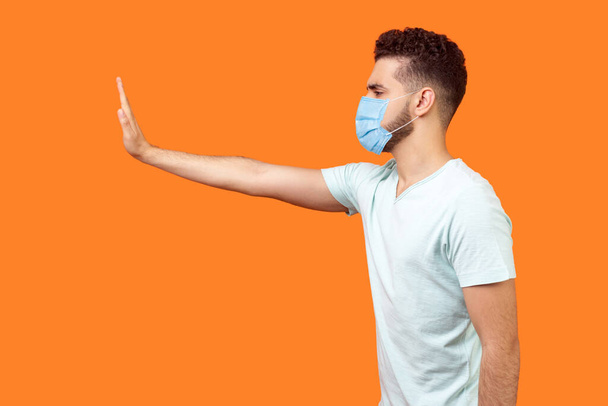 Stop, don't come any closer. Side view of serious bossy man with surgical medical mask standing holding out hand to left, prohibition gesture, saying no. studio shot isolated on orange background - Fotó, kép