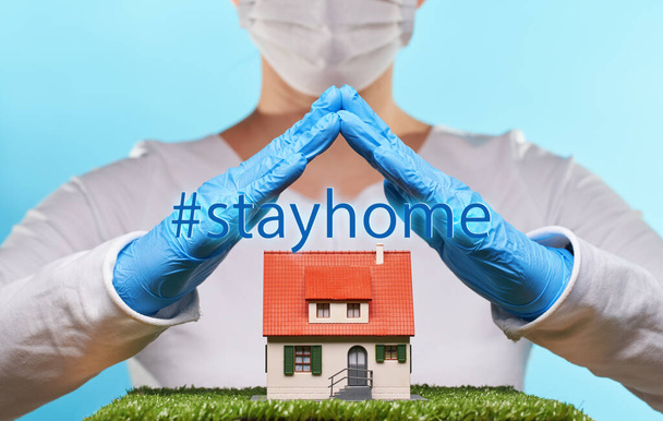 Γυναίκα με ιατρικά γάντια και μάσκα κρατά το χέρι πάνω από ένα σπίτι. Έννοια της διαμονής στο σπίτι, καραντίνα, ασφάλεια μέσα στο σπίτι. - Φωτογραφία, εικόνα