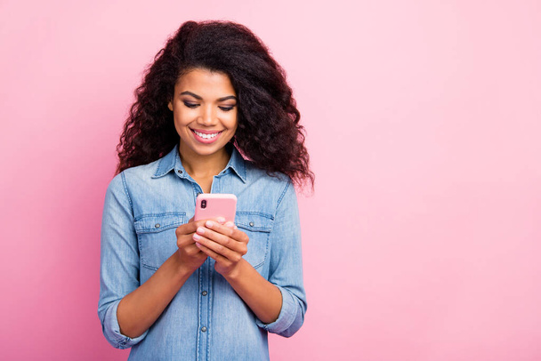 Portrait of positive cheerful afro american girl use her smartphone read social network news enjoy fun wear trendy outfit isolated over pink color background - Foto, imagen