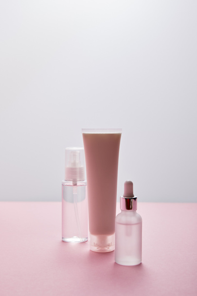 Cosmetics set of cream tube, spray with liquid and serum bottle on pink and grey background - Fotoğraf, Görsel