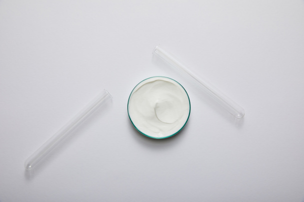 Bovenaanzicht van laboratoriumglaswerk met witte cosmetische crème en reageerbuizen op grijze ondergrond - Foto, afbeelding