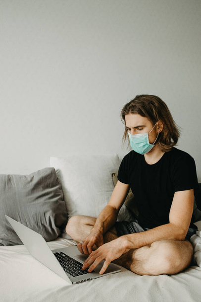 A white hipster man using laptop at home during quarantine at home. Caucasian man working remotely at home and wearing face mask for virus prevention.. Coronavirus concept.  - Foto, Imagen