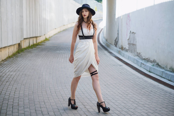 Νεαρή όμορφη σέξι γυναίκα που φοράει μοντέρνα ρούχα, λευκό φόρεμα, μαύρο καπέλο και δερμάτινη ξιφολόγχη. Μελαχρινή με μακριά μαλλιά ποζάρουν στο δρόμο της πόλης μια ηλιόλουστη μέρα. Εξωτερική φωτογραφία μόδας. - Φωτογραφία, εικόνα