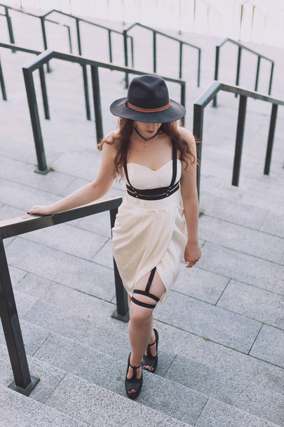Nuori kaunis seksikäs nainen yllään trendikäs asu, valkoinen mekko, musta hattu ja nahka miekkavyö. Pitkätukkainen brunette poseeraa kaupungin kadulla aurinkoisena päivänä. Outdoor muoti valokuvaus
. - Valokuva, kuva
