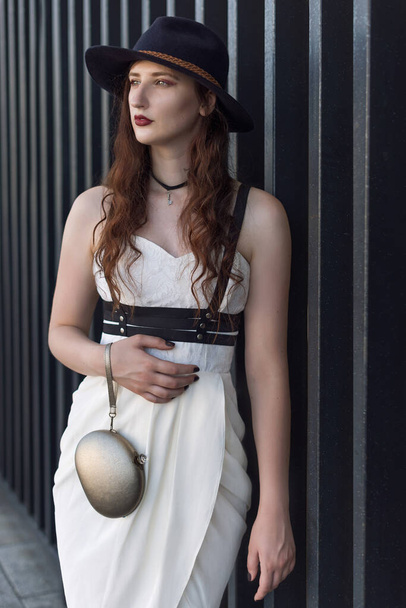 Fiatal gyönyörű szexi nő visel divatos ruhát, fehér ruha, fekete kalap és bőr kardöv. Hosszú hajú barna pózol a városi utcában. Kültéri divat fotózás. - Fotó, kép